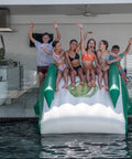 A group of six people posing on an inflatable pool slide with a crocodile face, positioned over a swimming pool in front of a modern house patio.