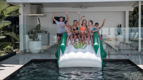 A group of six people posing on an inflatable pool slide with a crocodile face, positioned over a swimming pool in front of a modern house patio.