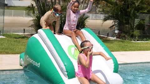 Children playing on a green and white inflatable slide in a pool with palm trees and a house in the background.